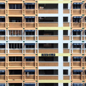 Op de afbeelding: Een foto van een meergezinswoning met een herhalend patroon van ramen, balkons en airconditioning-units. De gevel van het gebouw is een mix van beige, wit en blauw, met een bord met de tekst "564".
