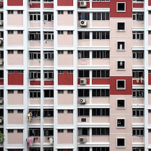 May include: A multi-story apartment building with a repeating pattern of windows and red and pink brick. Air conditioning units are mounted on the exterior walls. Some windows have laundry hanging outside. A green tree is visible in the lower left corner.