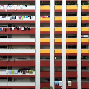 May include: A multi-story apartment building with a red, white, and yellow facade. Clothes are hanging on balconies. The building has a repeating pattern of windows and balconies, with laundry hanging on the balconies.