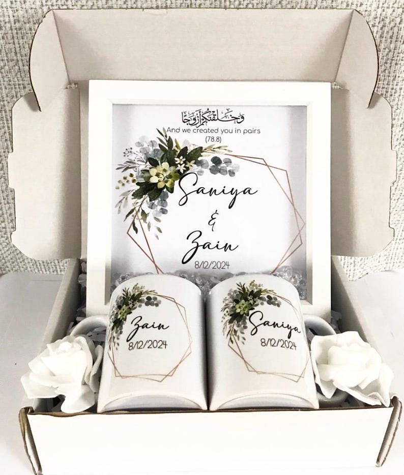 May include: A white gift box with a framed print featuring a floral design and the names "Saniya & Zain" with the date "8/12/2024". The box also contains two white mugs with the same floral design and names, and a white rose.