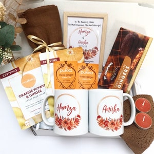 Könnte beinhalten: Ein Geschenkkorb mit einem braunen Schal, zwei Tassen mit den Namen "Hamza" und "Anisha" in goldenen Lettern, zwei Packungen Pukka Three Ginger Tee, eine Schachtel St. Andrews Dark Chocolate Ginger Biscuits, eine Gesichtsmaske und zwei Teelichter.