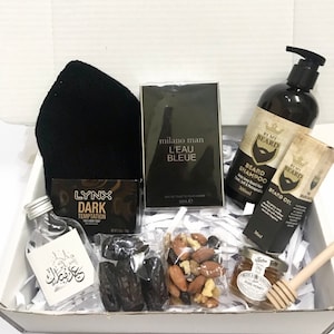 May include: A gift box filled with various items, including a black beanie, a bottle of cologne, a bottle of beard oil, a bottle of beard shampoo, a bar of soap, a small bottle of liquid, a bag of mixed nuts, dates, and a jar of honey. The box is lined with white shredded paper.