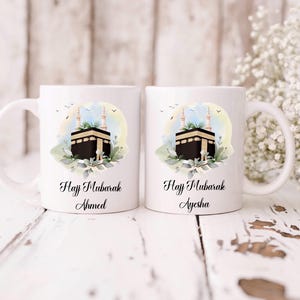 Może przedstawiać: Dwa białe ceramiczne kubki z uchwytem, każdy z kolorową ilustracją Kaaby w Mekce, z napisem "Hajj Mubarak" i imieniem. Kubki są ozdobione elementami kwiatowymi i małymi ptaszkami.