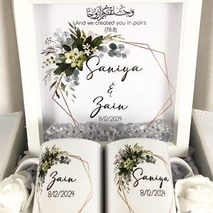 May include: Two white ceramic mugs with floral designs and gold geometric frames. The mugs are personalized with the names "Saniya" and "Zain" and the date "8/12/2024". A white frame with a floral design and gold geometric frame contains the Arabic text "وخلقتكم ازوجا" and the English text "And we created you in pairs (78.8)". The frame also includes the names "Saniya" and "Zain" and the date "8/12/2024".