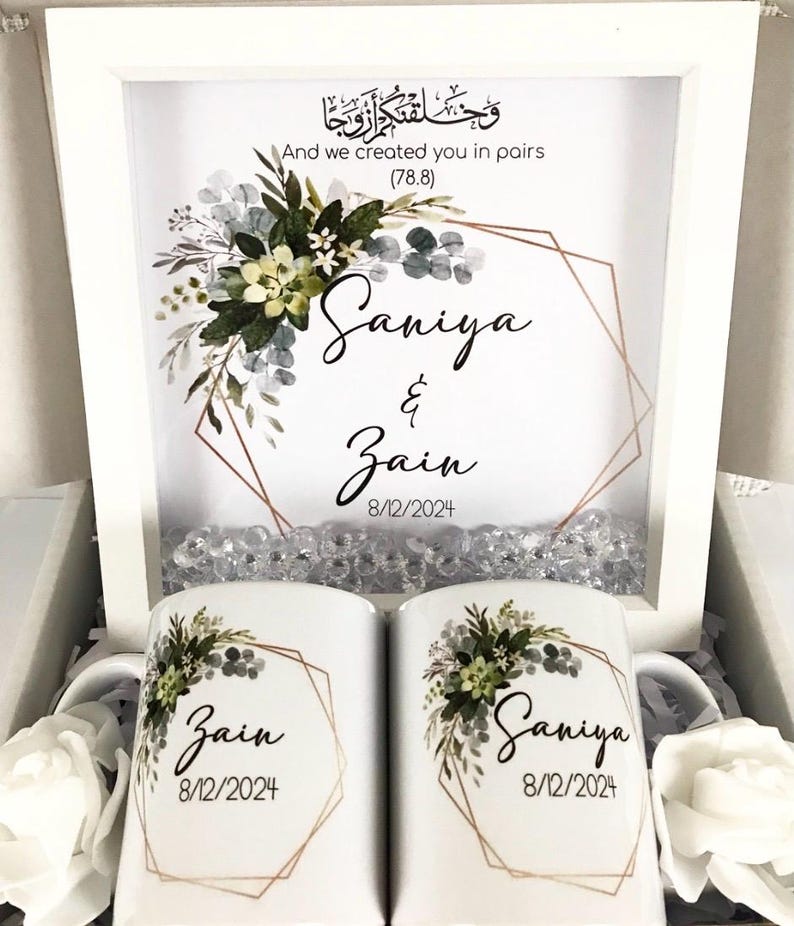 May include: Two white ceramic mugs with floral designs and gold geometric frames. The mugs are personalized with the names "Saniya" and "Zain" and the date "8/12/2024". A white frame with a floral design and gold geometric frame contains the Arabic text "وخلقتكم ازوجا" and the English text "And we created you in pairs (78.8)". The frame also includes the names "Saniya" and "Zain" and the date "8/12/2024".
