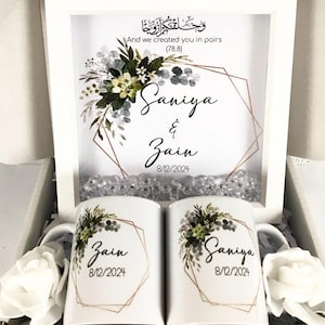 May include: Two white mugs with floral designs and gold accents. The mugs are personalized with the names "Saniya" and "Zain" and the date "8/12/2024". The mugs are in a white gift box with a framed print that says "And we created you in pairs (78.8)" with a floral design and gold accents. The print also includes the names "Saniya" and "Zain" and the date "8/12/2024".