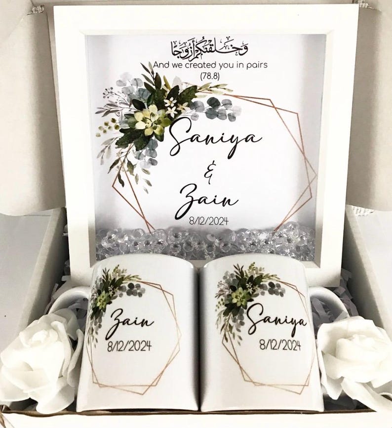 May include: Two white mugs with floral designs and gold accents. The mugs are personalized with the names "Saniya" and "Zain" and the date "8/12/2024". The mugs are in a white gift box with a framed print that says "And we created you in pairs (78.8)" with a floral design and gold accents. The print also includes the names "Saniya" and "Zain" and the date "8/12/2024".