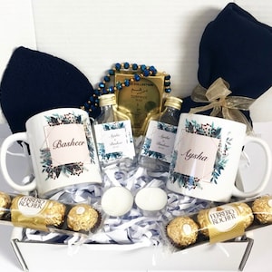 Könnte beinhalten: Eine weiße Geschenkbox gefüllt mit verschiedenen Artikeln, darunter zwei personalisierte Tassen, ein blauer Schal, zwei kleine Parfümfläschchen, goldene Gebetsperlen, eine goldene Schachtel mit arabischem Text und eine Schachtel Pralinen. Die Tassen haben Blumendesigns und die Namen "Basheer" und "Aysha" sind darauf geschrieben.