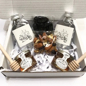 以下が含まれることがあります： アラビア語のラベルが付いた小さなガラス瓶2本、蜂蜜の小さな瓶2本、ミックスナッツの袋、ドライフルーツなど、さまざまなアイテムが入った白いギフトボックス。箱は白いシュレッダーペーパーで満たされています。