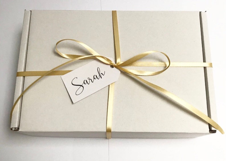 May include: A white cardboard box with a gold ribbon tied in a bow. A white tag with the name "Sarah" is attached to the ribbon.