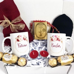 Könnte beinhalten: Eine weiße Geschenkbox gefüllt mit einem roten Schal, zwei weißen Tassen mit den Namen "Fatima" und "Bashir" darauf, einer kleinen Parfümdose, einer roten Perlenkette, zwei roten Kerzen und einer Schachtel Pralinen.