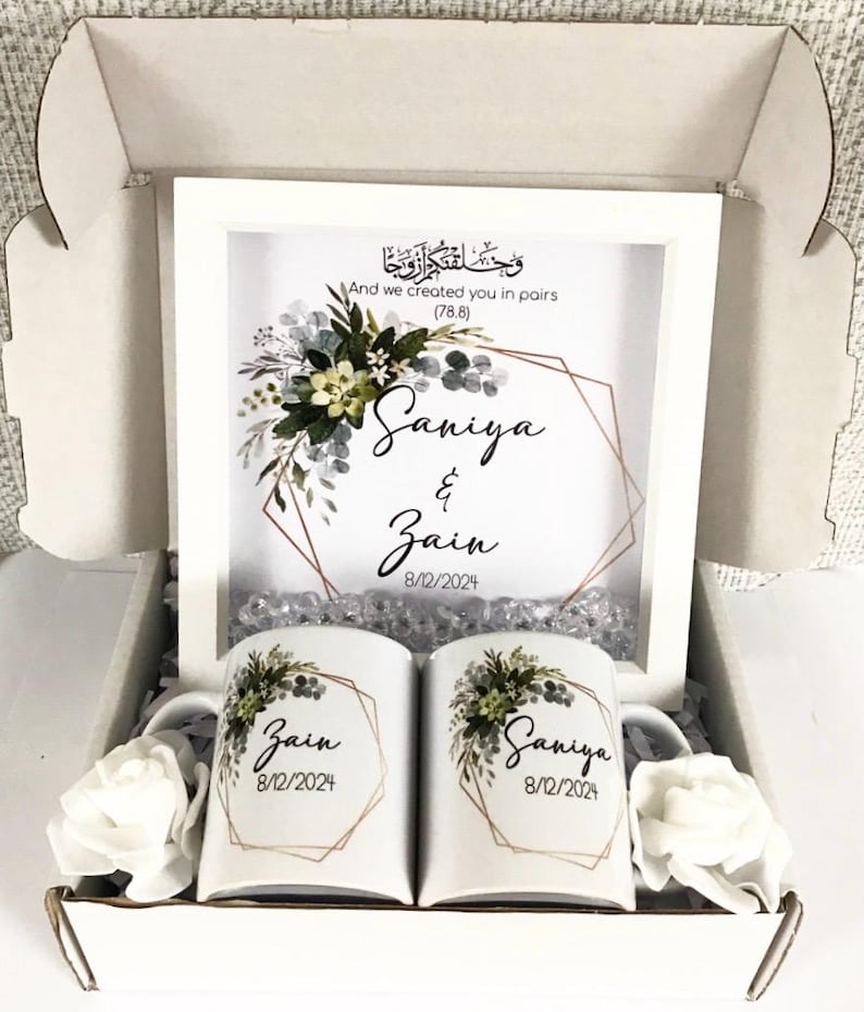 May include: A white gift box with a white frame containing a floral design and the names "Saniya & Zain" with the date "8/12/2024". The box also contains two white mugs with the same floral design and names, one with "Zain" and the other with "Saniya".