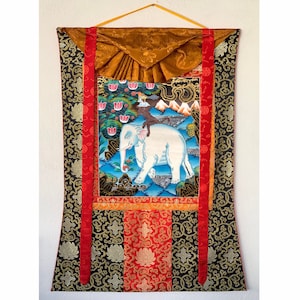 以下が含まれることがあります： 蓮の花が咲く風景の中を歩く白い象を描いた伝統的なチベットのタンカ画。タンカは金と赤の縁取りで額装され、金と銀の模様が施された黒い布地に張られています。