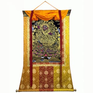 May include: A traditional Tibetan thangka painting depicting a golden deity with multiple arms and eyes. The deity is surrounded by intricate details and symbols, and the thangka is framed with a gold and red border.
