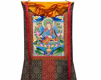 Puede incluir: Un tapiz thangka vertical que presenta una figura de deidad pintada en el centro, sentada sobre un trono de loto y rodeada por un paisaje vibrante. La obra está enmarcada por bordes de tela roja y dorada, rematada con una tela de seda amarilla.