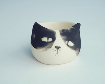 May include: A white ceramic planter shaped like a cat with black painted features. The cat has a sad expression with large eyes and a small nose.