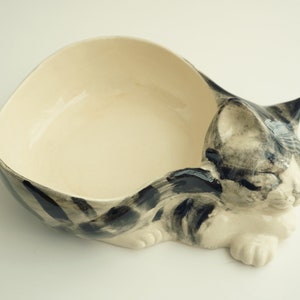 May include: A ceramic cat-shaped bowl with a white interior and black and gray stripes. The cat is curled up in a sleeping position.
