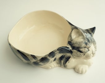 May include: A ceramic cat-shaped bowl with a white interior and black and gray stripes. The cat is curled up in a sleeping position.