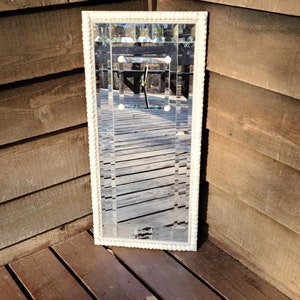 May include: A white framed mirror with a beaded border. The mirror is made of small, rectangular pieces of glass that create a geometric pattern. The mirror is reflecting a wooden deck and trees.
