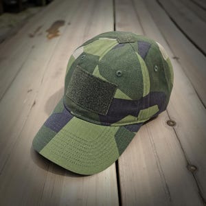 Peut inclure: Une casquette de baseball avec un motif camouflage vert, noir et beige. La casquette comprend un panneau auto-agrippant rectangulaire sur la couronne avant, des œillets cousus pour la ventilation et une visière incurvée avec des coutures.