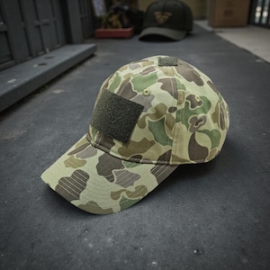 Könnte beinhalten: Eine Camouflage-Baseballkappe mit einem Muster in Grün-, Braun- und Beigetönen. Sie hat einen dunkelgrünen quadratischen Aufnäher auf der Vorderseite und einen kleineren oben. Der Schirm der Kappe ist gebogen. Das Muster ist Camouflage.