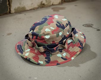 May include: A camouflage bucket hat featuring a multi-colored pattern of tan, dark blue, red, and light green shapes. The hat includes a wide brim, a stitched crown, and a fabric band with loops encircling the base, accented by small metal rivets.