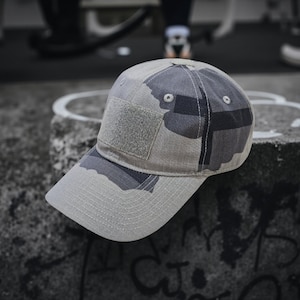 Könnte beinhalten: Grau-olivgrüne Camouflage-Baseballkappe. Die Kappe hat einen quadratischen Aufnäher auf der Vorderseite und einen gebogenen Schirm. Hergestellt aus strapazierfähigem Material mit einem strukturierten Design.