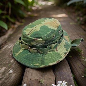 May include: A camouflage bucket hat featuring a green, brown, and tan woodland pattern. The hat includes a wrap-around fabric band with loops and two small silver-toned metal ventilation eyelets on the side of the crown and a wide brim.