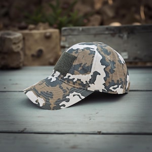 Puede incluir: Gorra de béisbol de camuflaje con un parche verde en la parte delantera. La gorra presenta un patrón de formas blancas, marrones y grises. La visera es blanca.