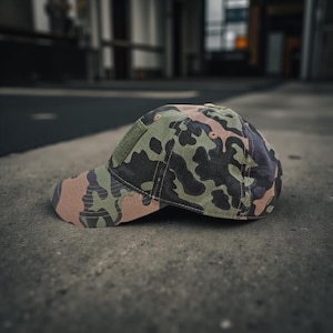 May include: Camouflage baseball cap with a green hook-and-loop patch. The cap has a pattern of green, brown, and black shapes. The brim is a solid brown colour. The cap is resting on a concrete surface.