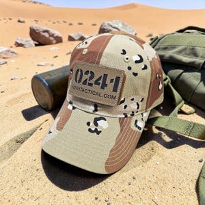 May include: A tan and brown camouflage baseball cap with a patch that reads "024-1" and "024TACTICAL.COM". The cap is set against a desert backdrop with a green bag and a dark green bottle.