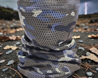 May include: A gray and blue camouflage pattern neck gaiter. The gaiter is made of a breathable, perforated fabric. The design features a mix of gray, blue, and white shapes. The gaiter is shown outdoors on a rocky surface.