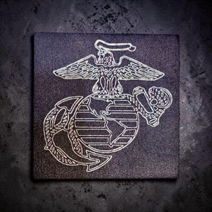 May include: Square, dark gray tile featuring a white outline of the United States Marine Corps emblem. The emblem includes an eagle, globe, and anchor design. The tile has a textured surface and is set against a dark background.