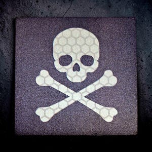 May include: Square, dark gray coaster featuring a white skull and crossbones design. The skull and bones have a honeycomb pattern. The coaster is a square shape.