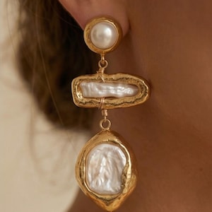 May include: Gold-coloured drop earrings with three sections. The top has a round pearl, followed by a rectangular pearl in a gold frame, and a larger, teardrop-shaped pearl at the bottom, also framed in gold. The earrings have a textured, organic design.