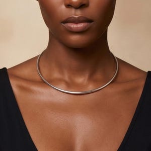 Könnte beinhalten: Eine polierte silberne Choker-Halskette. Die Halskette hat ein einfaches, glattes und abgerundetes Design. Das Model trägt ein schwarzes Oberteil. Der Hintergrund ist beige.