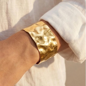 May include: A wide, hammered gold cuff bracelet. The textured surface reflects light, creating a shimmering effect. The bracelet is worn on a wrist, paired with a white long-sleeved shirt.