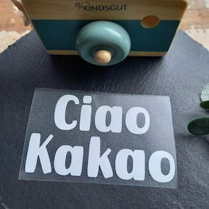 Könnte beinhalten: Eine Holzspielzeugkamera mit türkisfarbenen und natürlichen Holzakzenten, der Text "KINDSGUT" ist sichtbar. Ein klarer Aufkleber mit den Worten "Ciao Kakao" in weißen Blockbuchstaben befindet sich im Vordergrund.