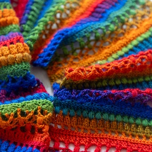Puede incluir: Una bufanda de crochet arcoíris con una variedad de colores, incluyendo rojo, naranja, amarillo, verde, azul, índigo y violeta. La bufanda está hecha con un punto de crochet que crea un aspecto texturizado.