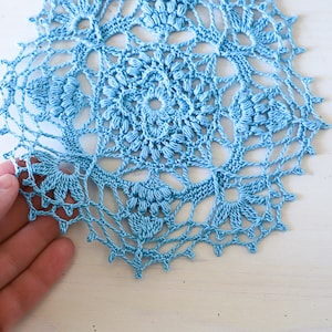 Könnte beinhalten: Eine zarte, blaue Häkeldeckchen mit einem runden Design. Das Deckchen hat eine Mitte mit einem kleineren Kreis und komplizierten Mustern, die nach außen strahlen.