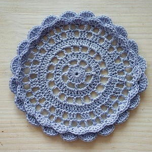 May include: A handmade, light blue crocheted doily with a scalloped edge. The circular design features an open, lacy pattern radiating from the center. This decorative piece is likely used for home decor.