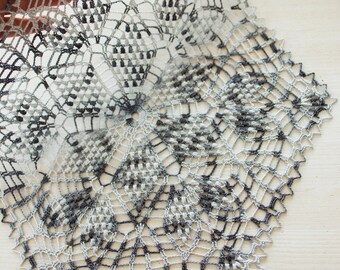 May include: A handmade, openwork doily in a circular pattern. The doily features a detailed, intricate design with a gradient of gray and black threads. The edges are scalloped, adding a decorative touch to this textile.