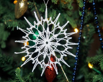 Puede incluir: Un adorno de copo de nieve blanco tejido a ganchillo cuelga de un árbol de Navidad. El copo de nieve tiene un diseño calado delicado con múltiples puntas. También se ven otros adornos y ristras de cuentas.
