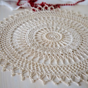May include: A delicate, off-white crocheted doily with a circular design. The doily has a centre circle with a smaller circle inside, and the design radiates outwards in a symmetrical pattern.