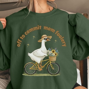 May include: Dark green sweatshirt with the phrase "off to commit mom foolery" in a circular design. The graphic features a goose wearing sunglasses and a hat, riding a yellow bicycle with a basket of sunflowers.