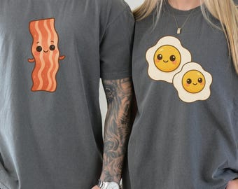 May include: Two charcoal gray t-shirts with matching food-themed graphics. One shirt features a cartoon bacon strip, the other has two cartoon fried eggs. The shirts are worn by two people holding hands.