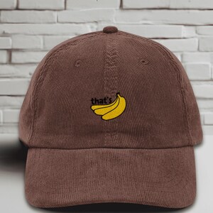 Brown baseball cap with a curved brim featuring an embroidered yellow banana design and the word “that’s” stitched in black above it. The hat is displayed upright against a white brick wall background, showcasing its soft, unstructured style and casual look.