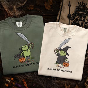 May include: Two crewneck t-shirts, one olive green and one cream, each featuring a cartoon frog illustration. The green shirt shows a frog in a cape with a sword and pumpkin bucket, text reads: WE PILLAGE CANDY AT DAWN. The cream shirt shows a frog in a witch hat with a sword and pumpkin bucket, text reads: WE CLAIM THE SWEET SPOILS.