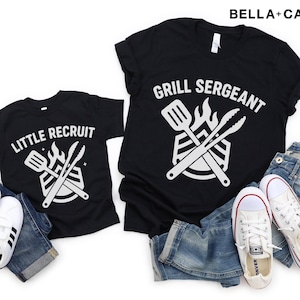 May include: Black t-shirts with white graphics and text. One shirt says "Grill Sergeant" and the other says "Little Recruit". The shirts are paired with blue jeans and white sneakers. A fun theme for family gatherings and barbecues.