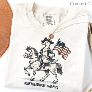May include: Cream-colored Comfort Colors t-shirt featuring a cartoon duck riding a horse and holding an American flag. The duck is dressed in colonial attire. The text "HONK FOR FREEDOM - 1776-2026" is printed below the graphic.